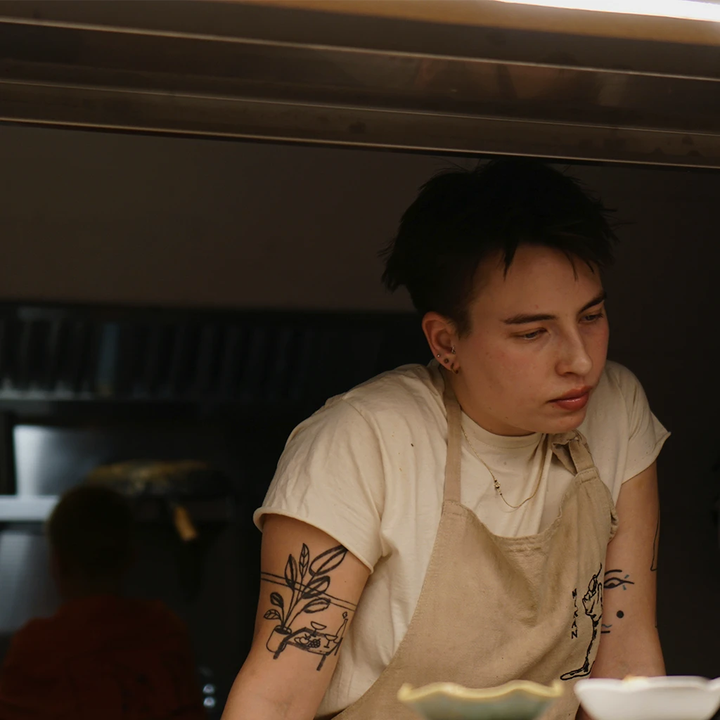 a white lady with short hair, peircings and tatoos,wearing an apron looking slightly sad