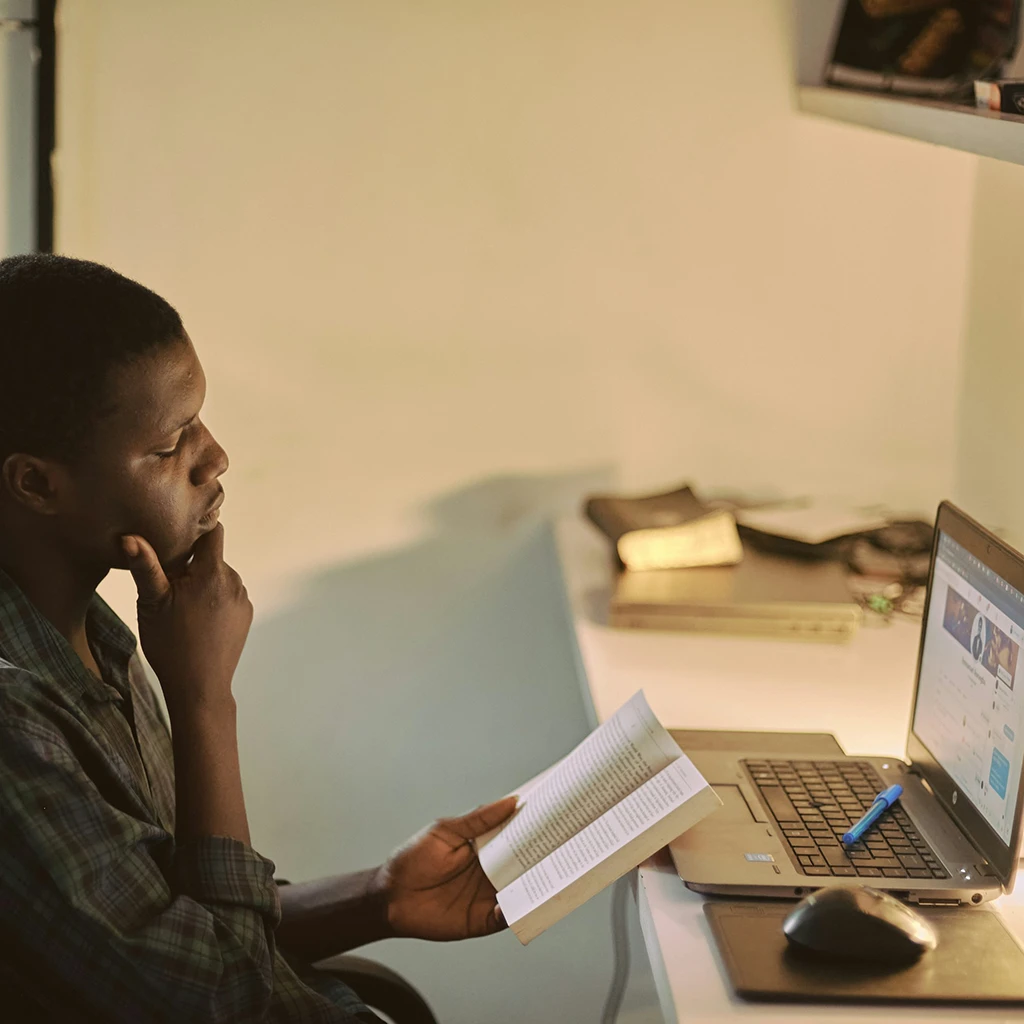 a black male holding his chin think as he reads a book infron of his laptop